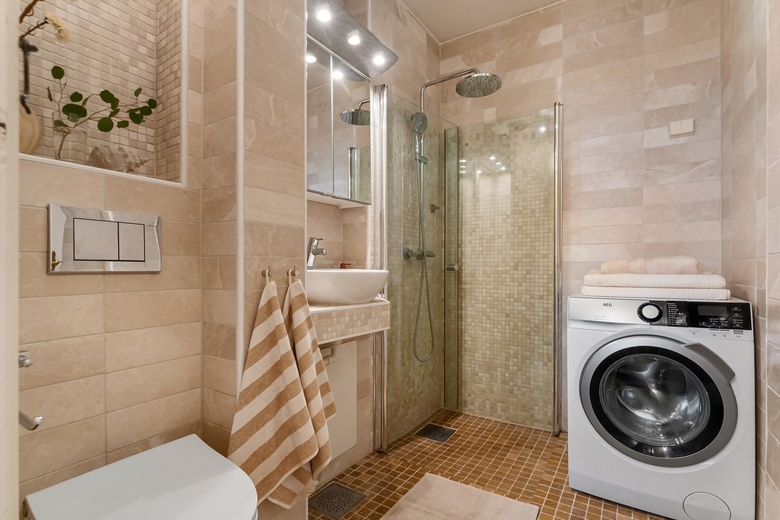 Baderommet er utstyrt med dusj på gulv med dører i herdet glass, veggfestet klosett, servant, opplegg for vaskemaskin og overskap med speilfronter- og belysning Galleribilde