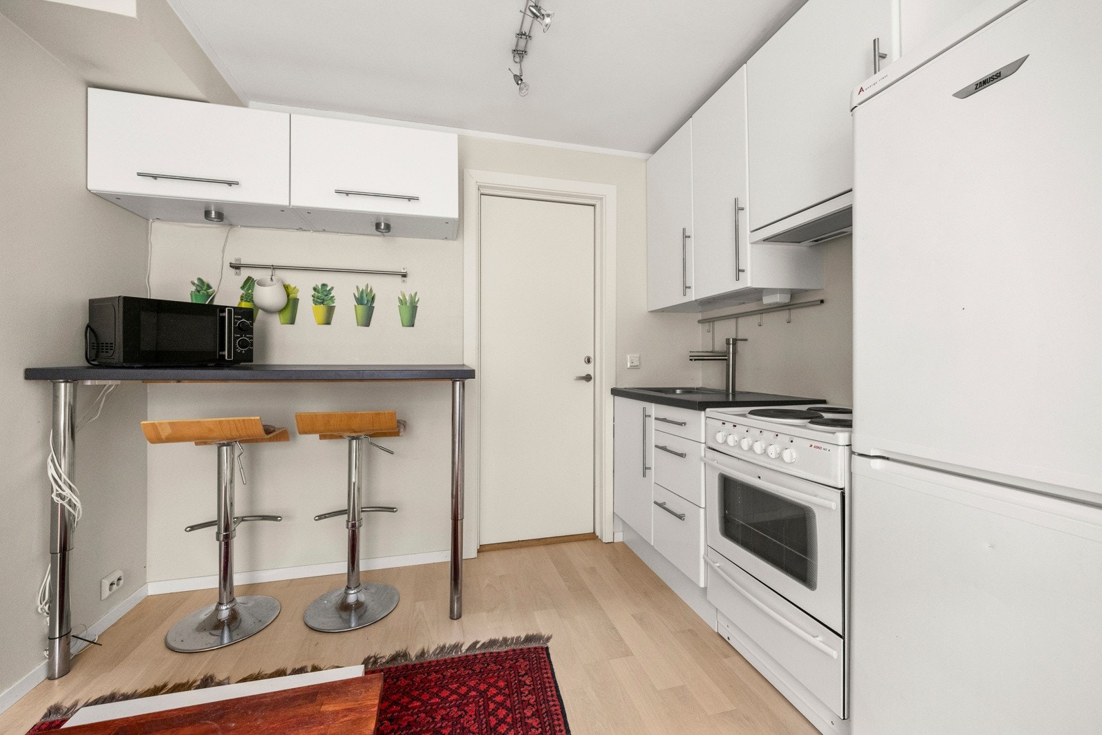 Hybeldelens kjøkken har en praktisk innredning med slette fronter, laminat benkeplate og en åpen løsning mot stuen, som skaper en sosial og funksjonell planløsning. Galleribilde