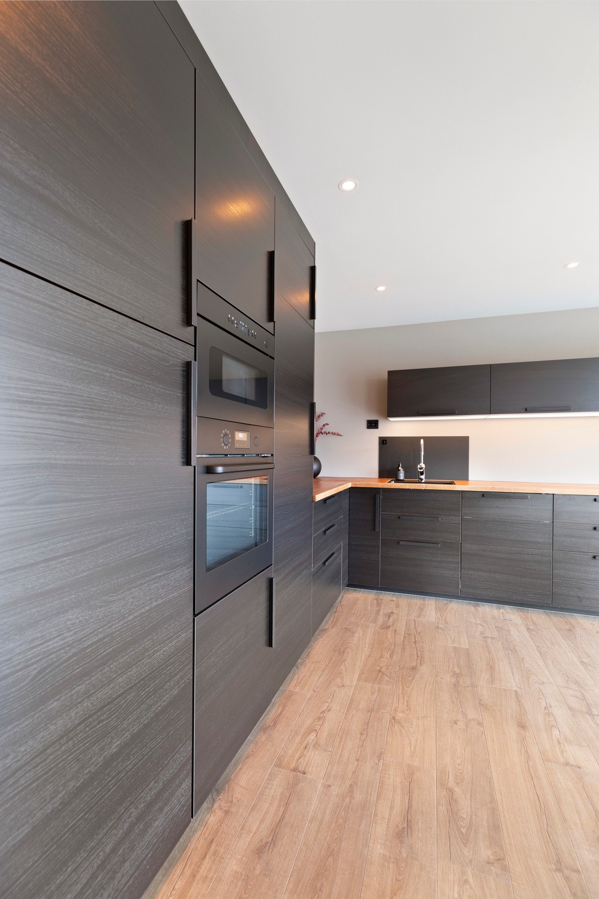 Kjøkkenet har montert en romslig, L-formet innredning med benkeskap, høyskap og overskap med glatte, mørke fronter Galleribilde