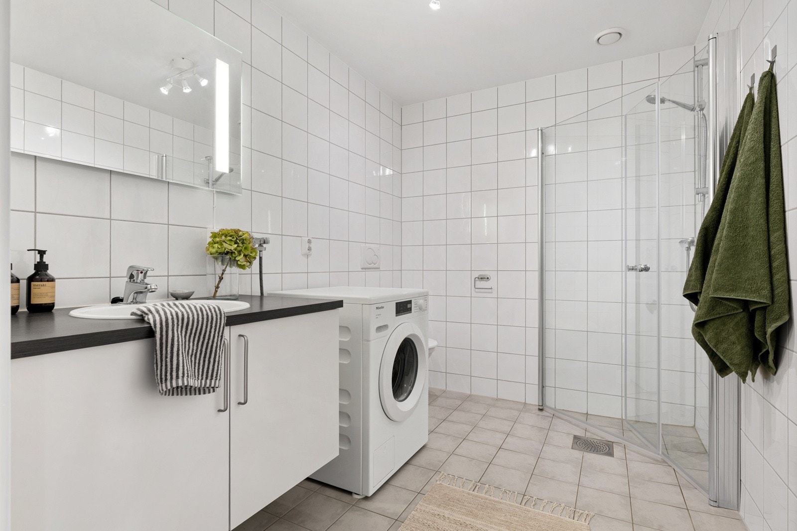 Det er god plass til oppbevaring av baderomsartikler i servantskap. Øvrig innredning består av speil med integrert belysning, en praktisk og åpen dusjløsning, vegghengt toalett og opplegg for vaskemaskin. Galleribilde