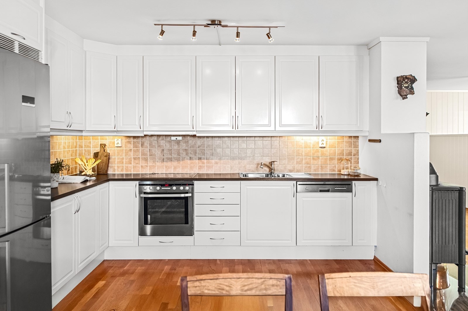 Kjøkkenet er tidløst og funksjonelt, utformet i en praktisk L-form med god skapplass og arbeidsflyt. Innredningen har hvite speildører og en lys laminatbenkeplate med dobbel oppvaskkum. Galleribilde