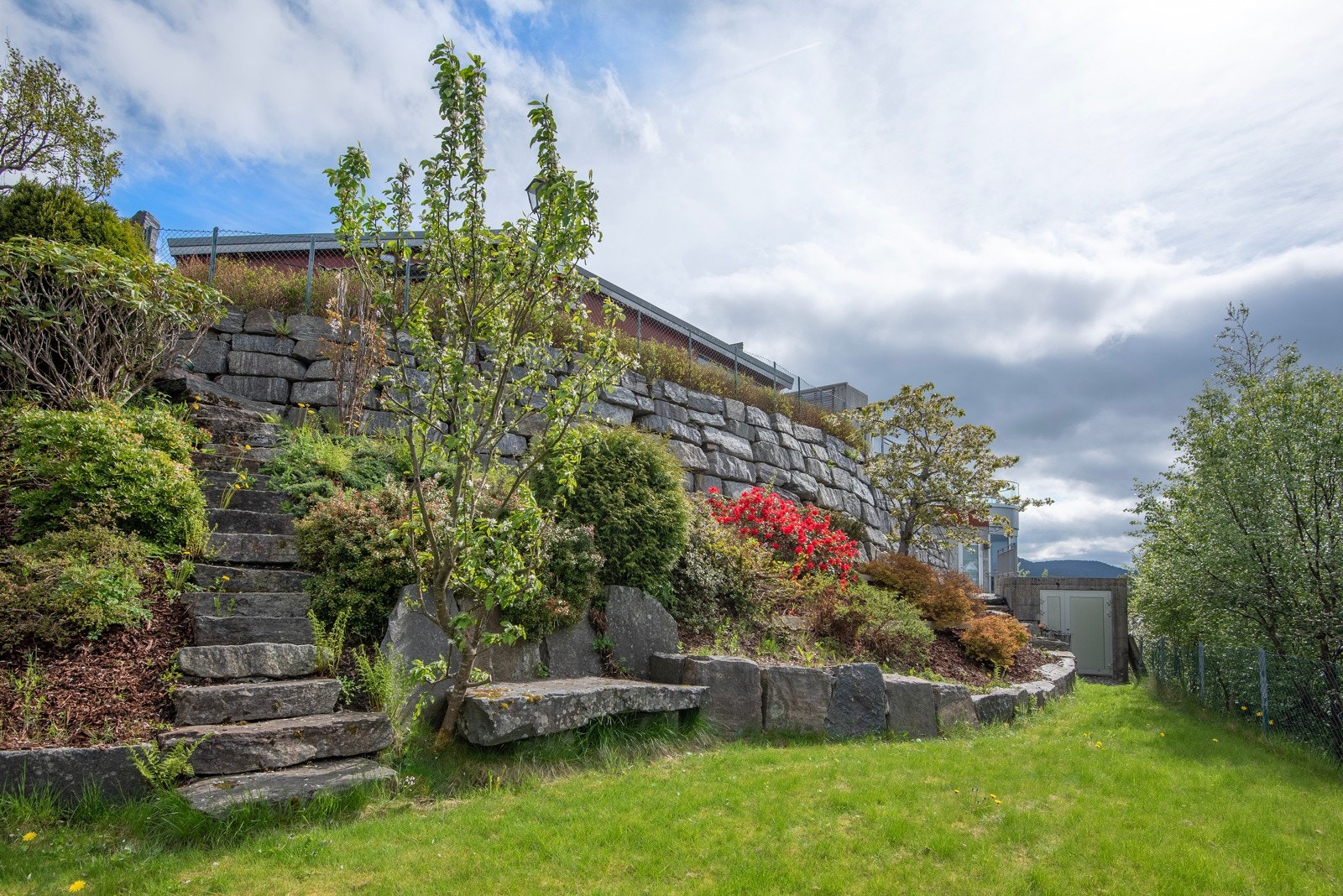 På del av tomten er det også hage med plen og beplantning og fine natursteinsmurer. Døren i høyre bildekant er en stor lagringsbod bygd i betong. Galleribilde