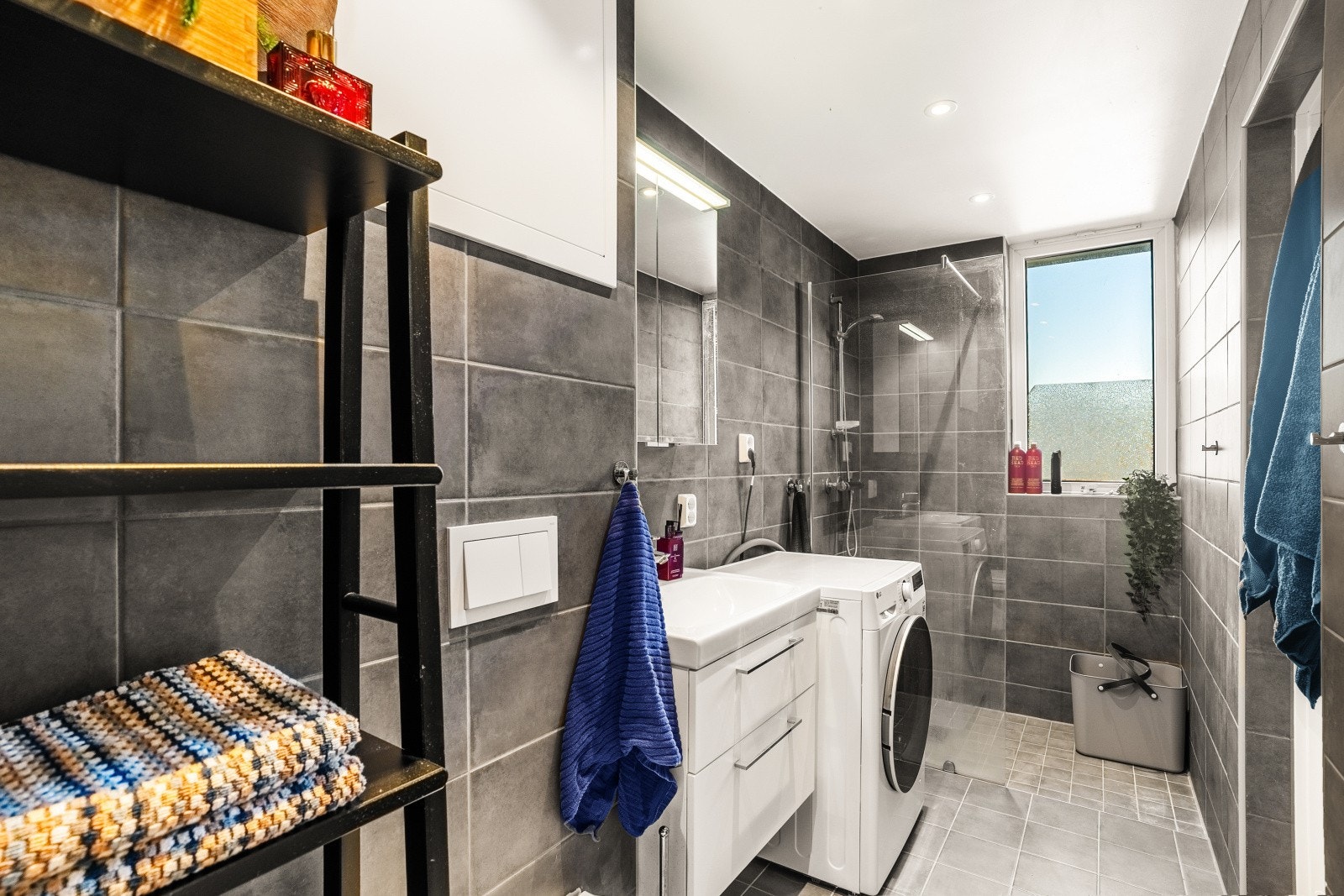 Moderne bad med dusjhjørne, vaskemaskin, servant og vindu som gir naturlig lys. Veggfliser i grått gir et stilrent uttrykk. Galleribilde