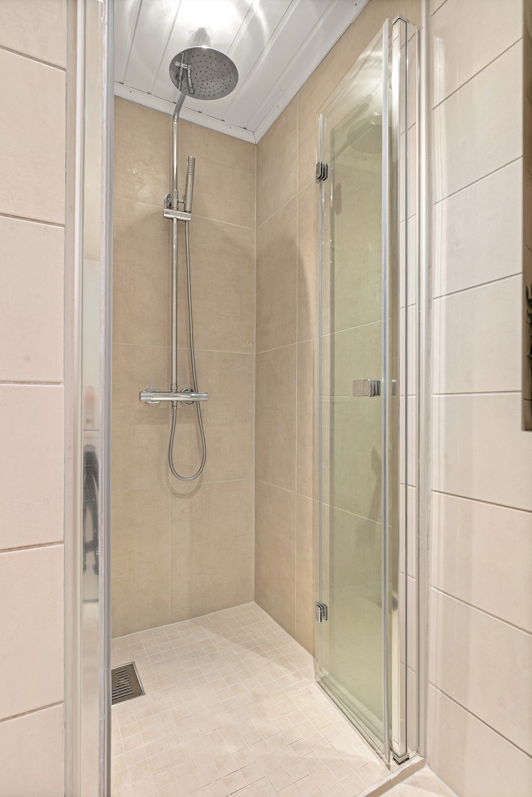 Et stilrent dusjhjørne er utstyrt med regnfallsdusj, innfellbare dører og store veggfliser i en moderne jordtone. Galleribilde