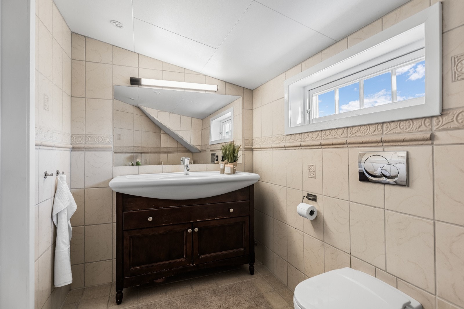 Badet oppe er innredet med beige fliser, vegghengt toalett og opplegg for badekar. Bderomsinnredningen består av servant med mørke skuffer og et speil med belysning. Galleribilde