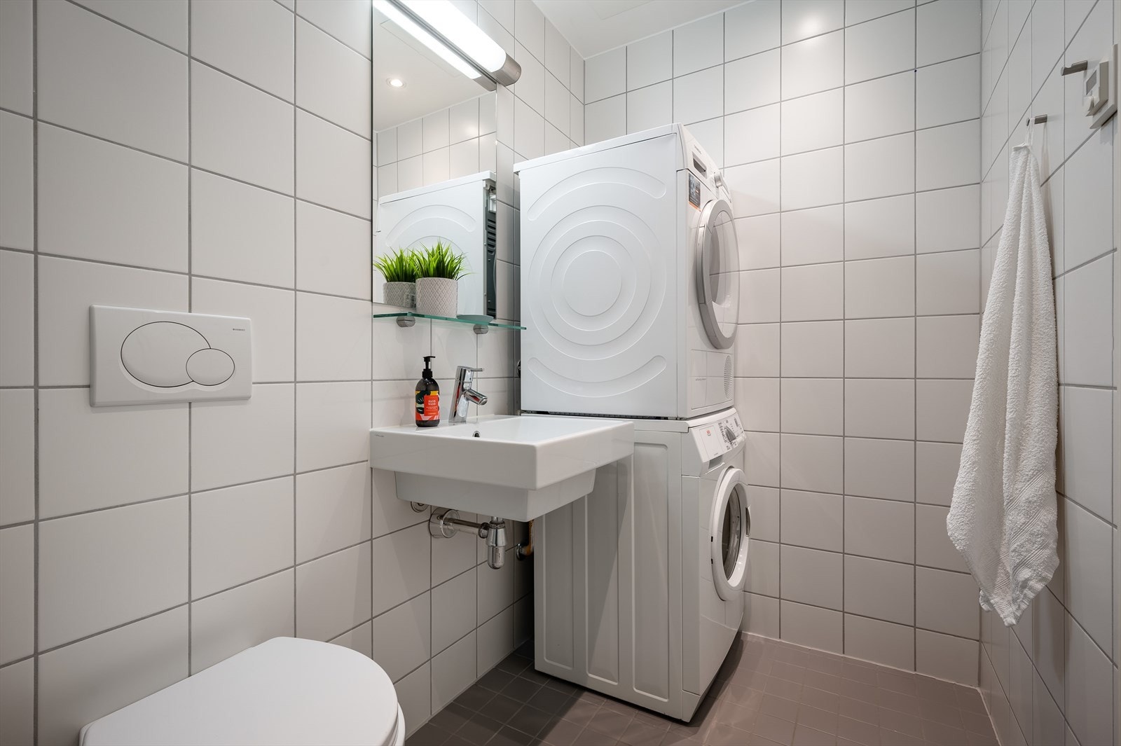 Baderommet inkluderer vegghengt toalett, servant med speil, dusjhjørne med glassvegg og forheng, opplegg for vaskemaskin og tørketrommel. Galleribilde