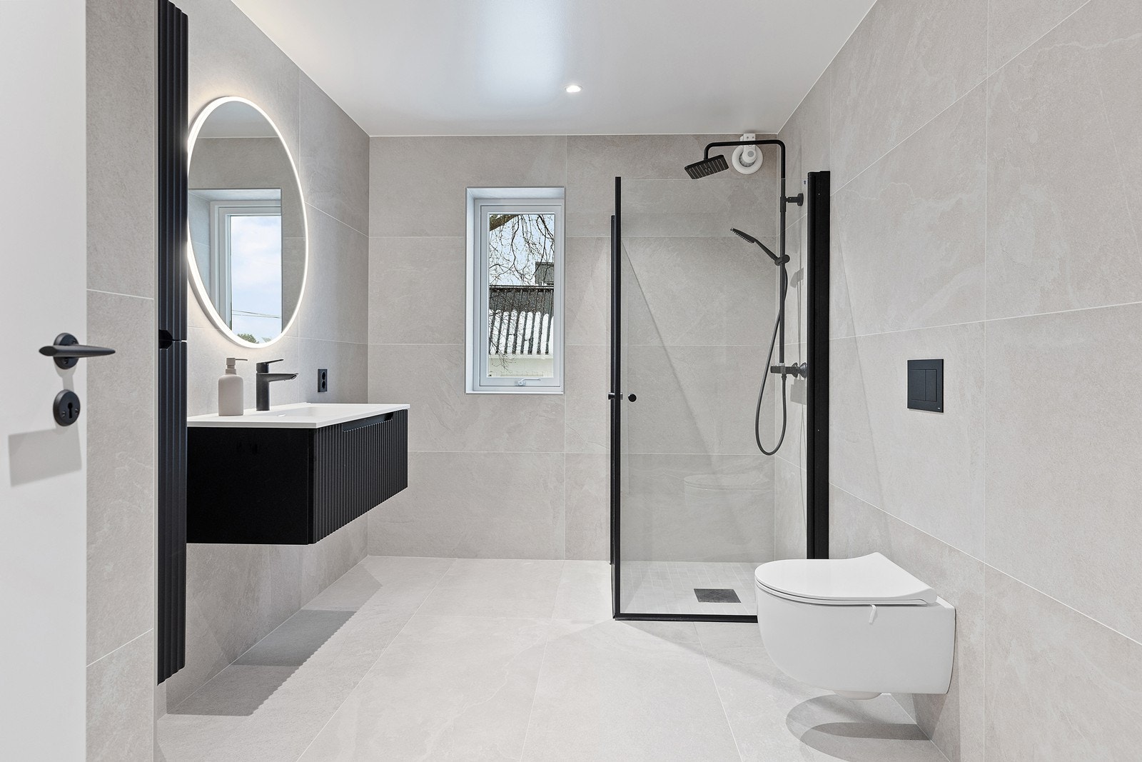 Baderommet har store fliser på gulv og vegger og varme i gulvet, vegghengt wc og regnfallsdusj Galleribilde