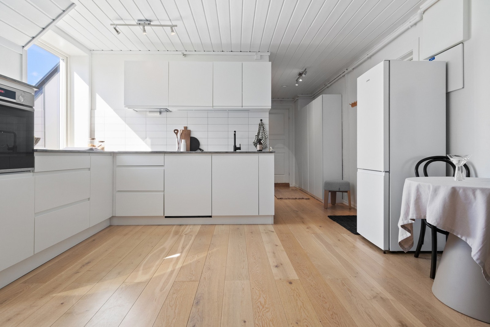 Innredningen har hvite, glatte fronter, hvite fliser over benk, benkebelysning og grå benkeplater med sort oppvaskkum. Galleribilde