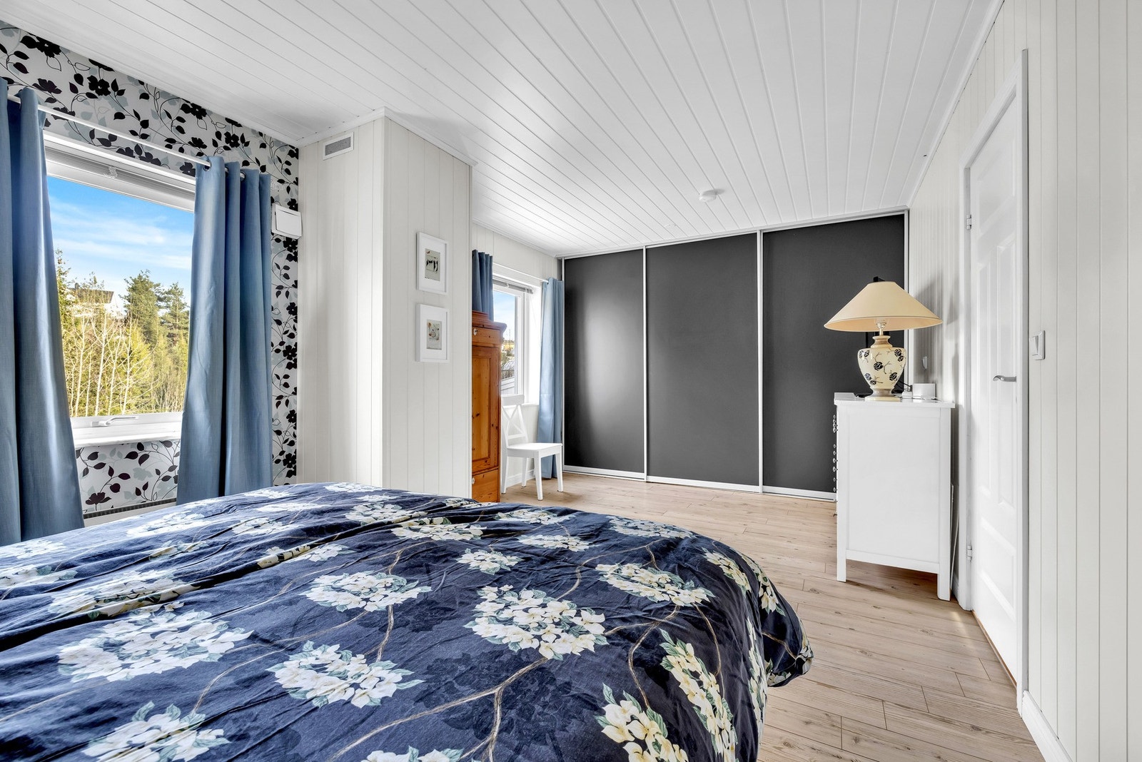 Hovedsoverommet har gulvflater av parkett, hvitmalte panelplater på vegger og himling, i tillegg til to kontrastvegger med tapet i blomstrete mønster. Her får du lysinnslipp gjennom to vindusflater som bidrar til å gi rommet en lys og luftig følelse. Galleribilde