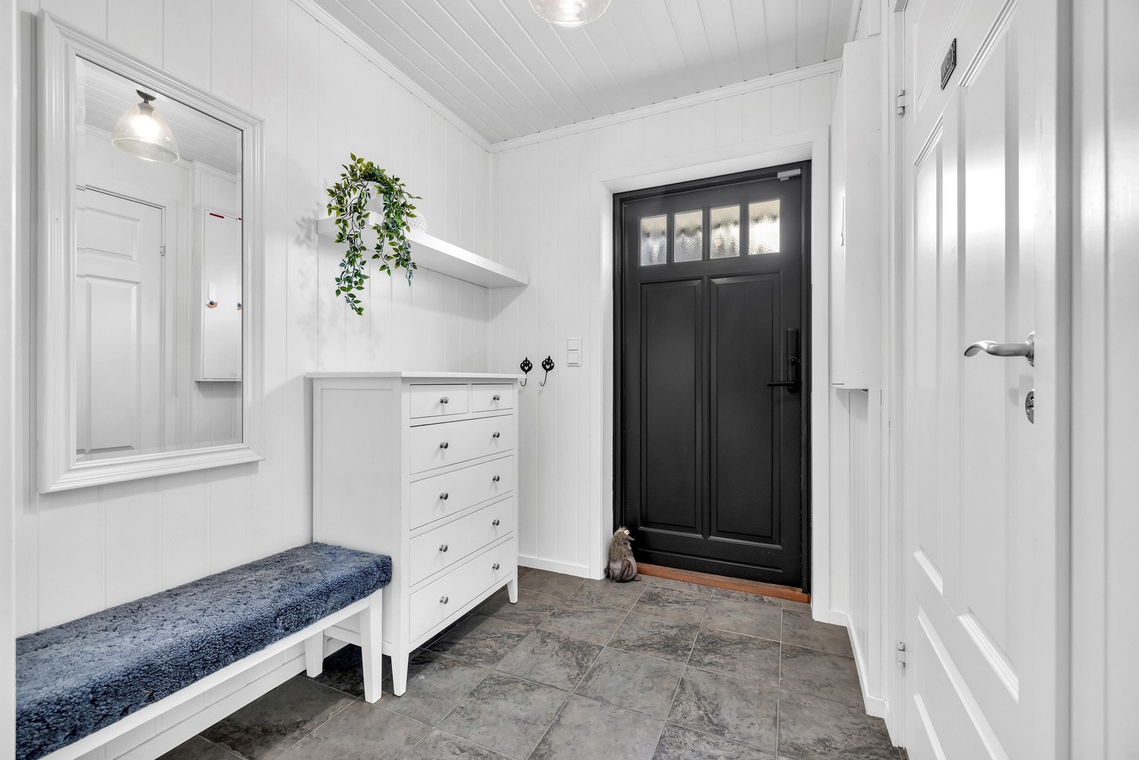 Velkommen inn i en lys entré med grå fliser på gulvet, hvitmalte panelplater på vegger og i himlingen. Her er det plass til en kommode for oppbevaring og en krakk man kan sitte på i mens man tar på seg sko. Fra entréen er det adkomst til et WC rom. Galleribilde