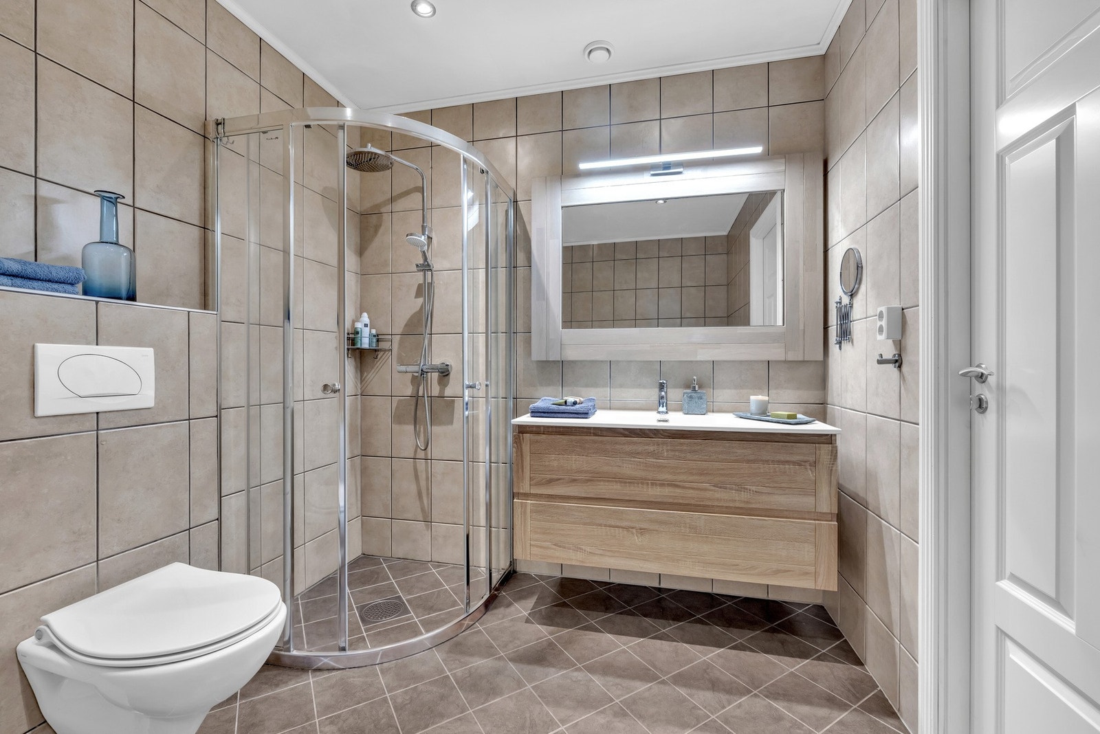 Her er det beige/brunt flislagt gulv med gulvvarme og vegger, samt hvitmalt himling med downlights. Baderommet har innredning med servant med ett-greps armatur, speil med belysning, vegghengt toalett, dusjhjørne med skyvedører og dusjhode samt hånddusj. Galleribilde