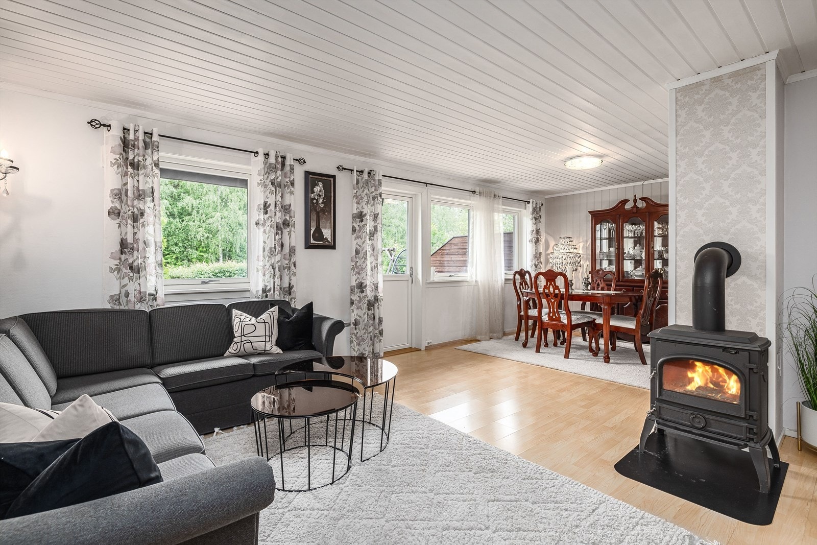 Stuen i vinkel har fine sosiale soner og flere møbleringsmuligheter. I dag er stuen innredet med romslig sofagruppe nær peisvedovn og en hyggelig spisestue. Galleribilde