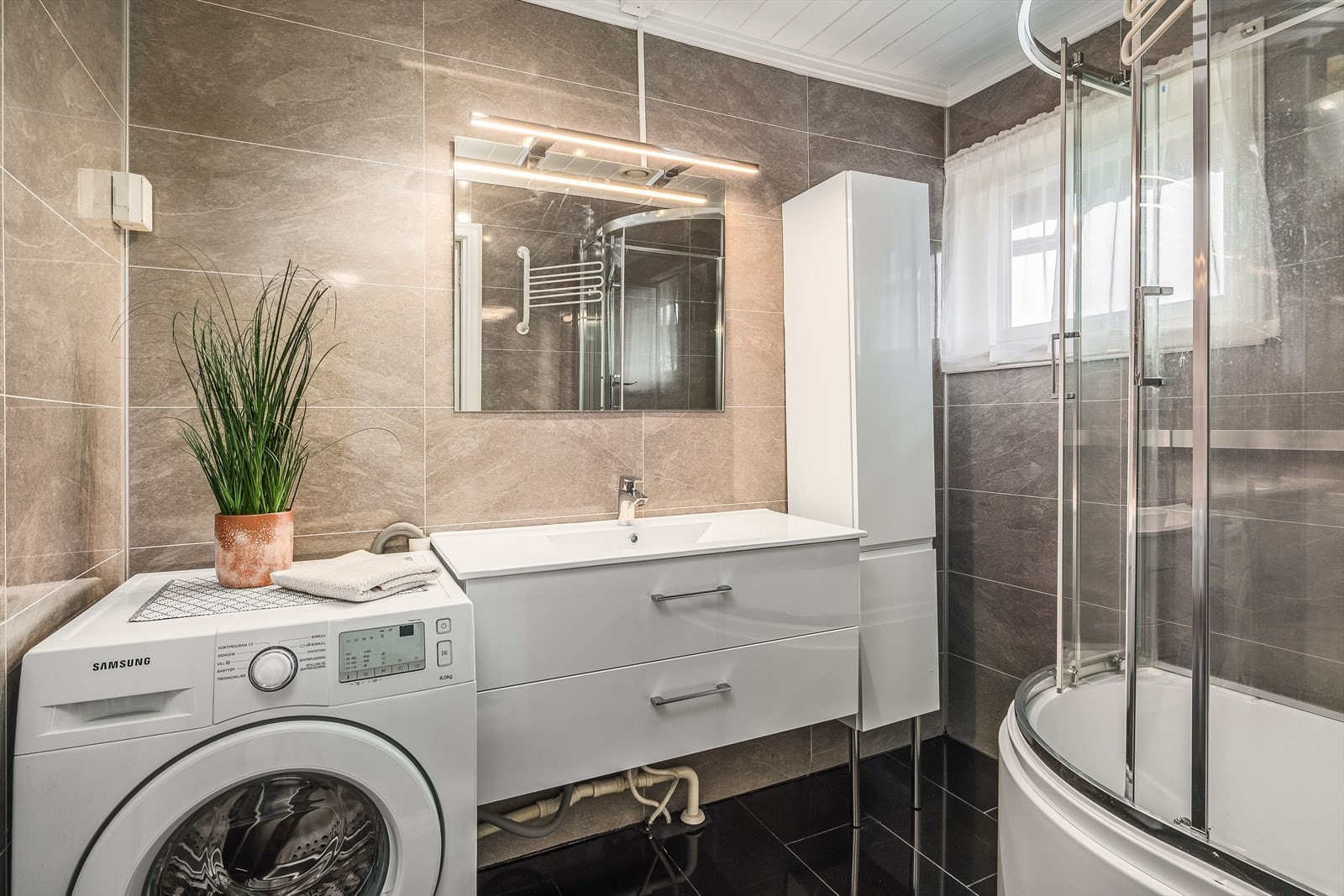 Badet har en lun fremtoning med flislagt gulv og baderomsplater med flisemønster på vegger. Nyere og moderne innredning med speil og belysning over. Galleribilde