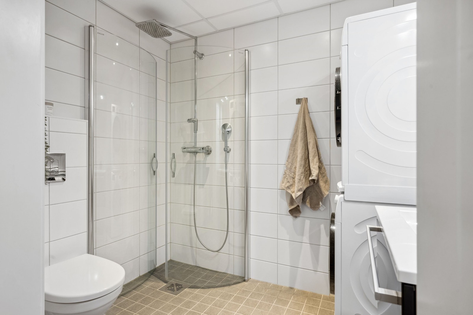 Flislagt gulv, dusjhjørne, vegghengt toalett og servant, og opplegg for vaskemaskin. Galleribilde