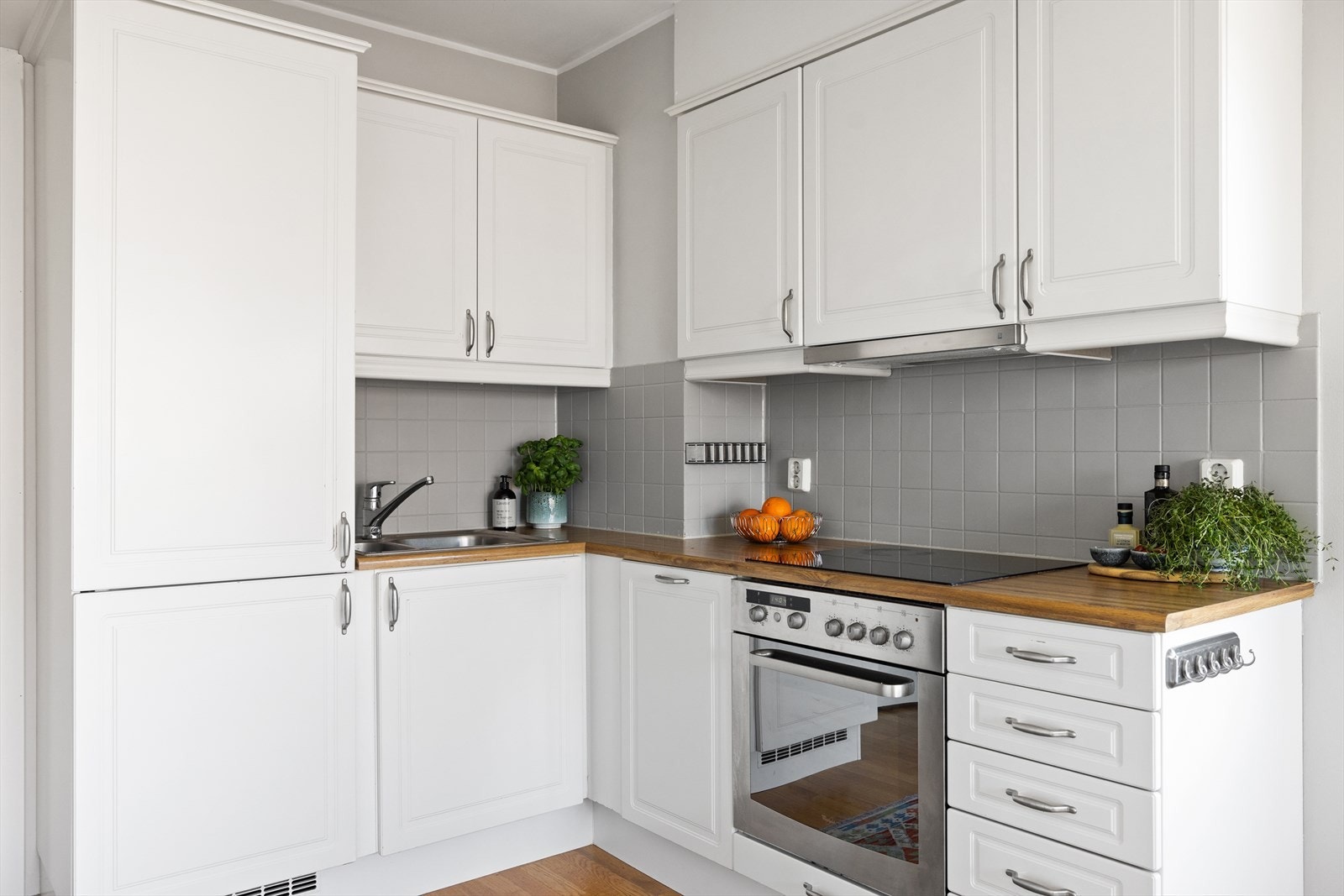 Innredningen har profilerte hvite fronter og laminat benkeplate med nedfelt kum. Det er montert belysning under overskapene og fliser på vegg mellom benkeplate og overskap. Galleribilde