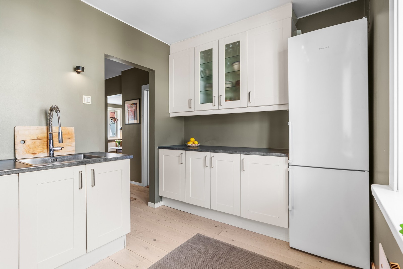 Ellers er det god plass for frittstående kjøl/fryseskap, og innredningen har takhøye veggskap, hvite rammefronter, vitrineskap, benkeplate i grå laminat og vaskekum i stål med spylekran. Galleribilde