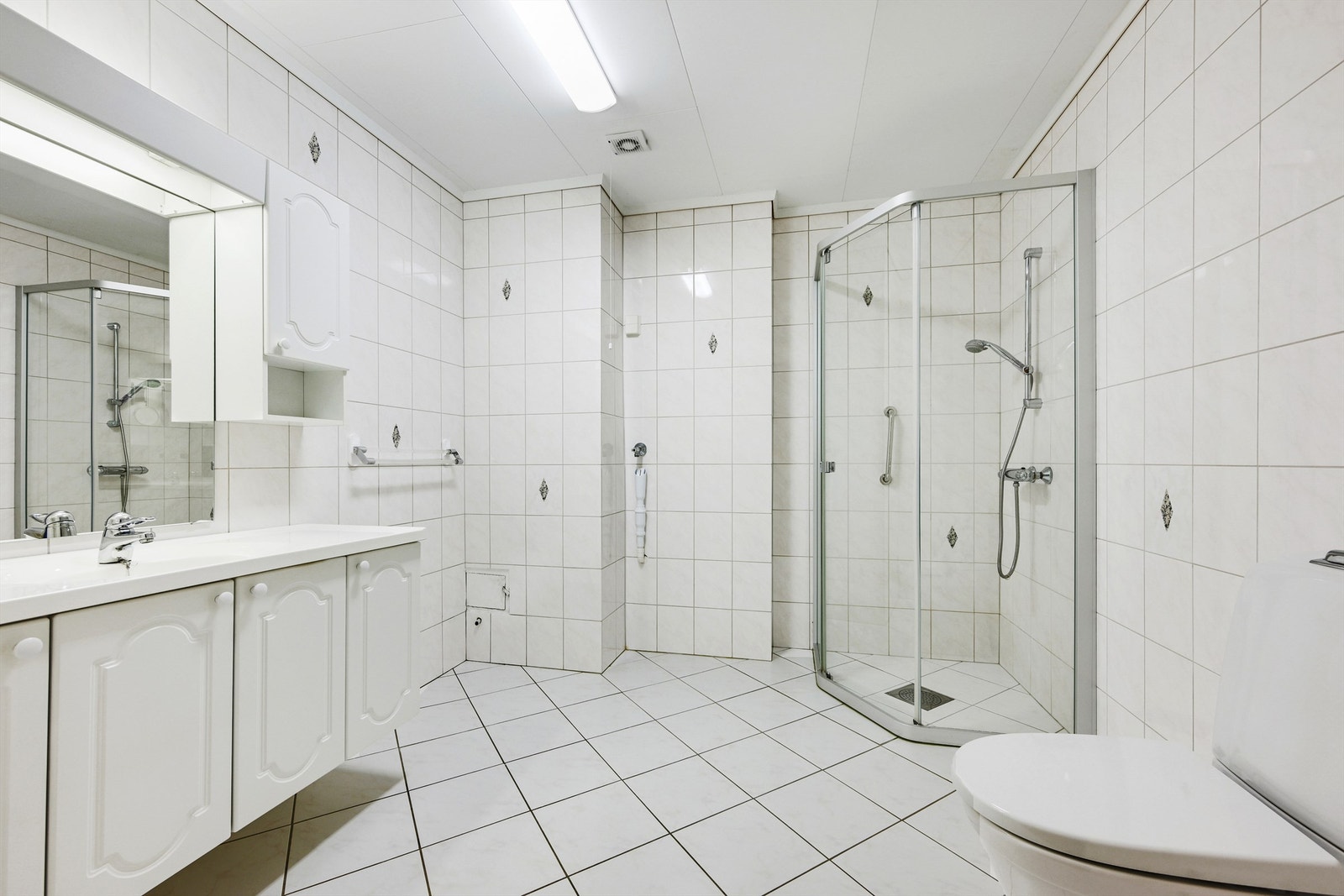 Badet er innredet med dusjdører, gulvmontert toalett, opplegg for vaskemaskin og innredning med nedfelt vask og speilskap over. Galleribilde