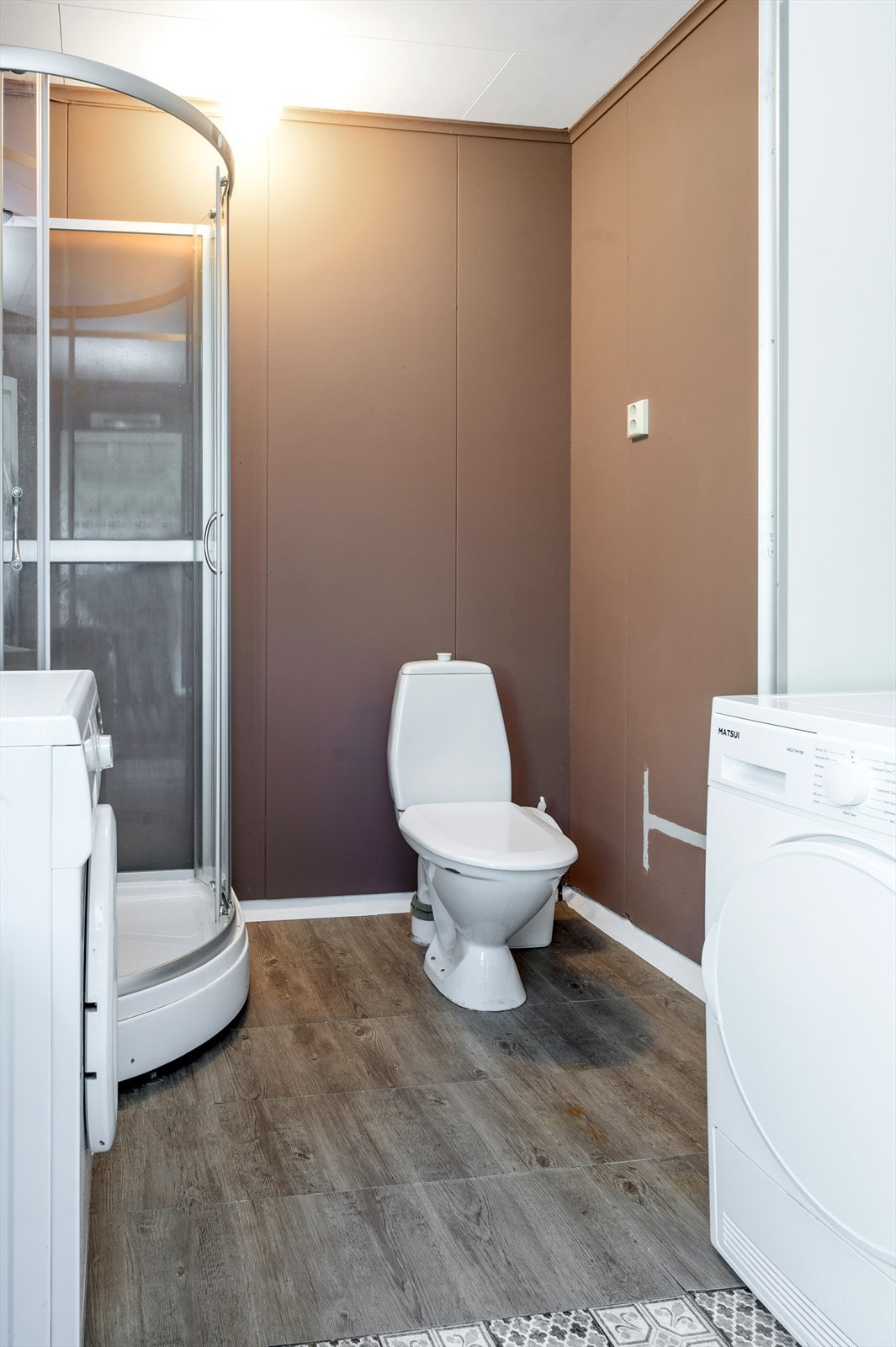 Bad/vaskerom med vinyl på gulv, enkelte løse laminatfliser og malte plater på vegger - Her finner du toalett, dusjkabinett, vegghengt skyllekar og opplegg for vaskemaskin Galleribilde