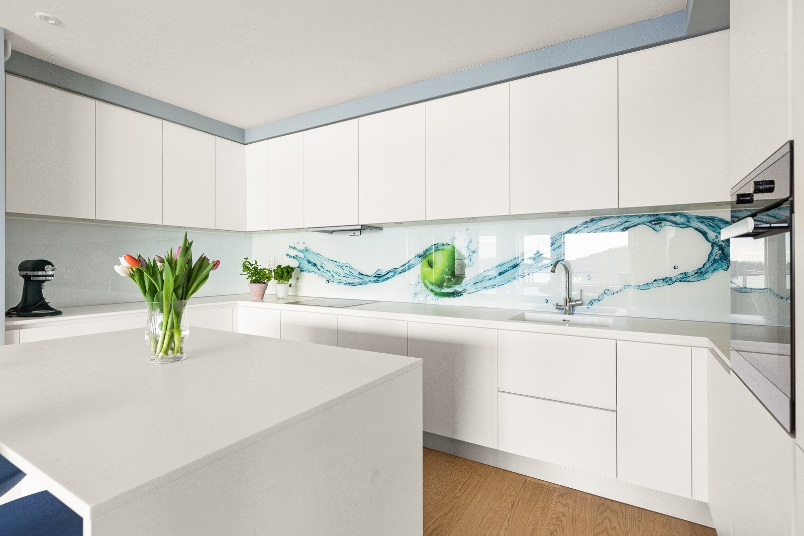 Innredning har hvite glatte fronter, lys benkeplate (Blanco Zeus 20 mm kompositt-sten) med helstøpt servant, glassplater på vegg over benk med motiv, downlights under overskap, avtrekksvifte Cube og komfyrvakt installert. Galleribilde