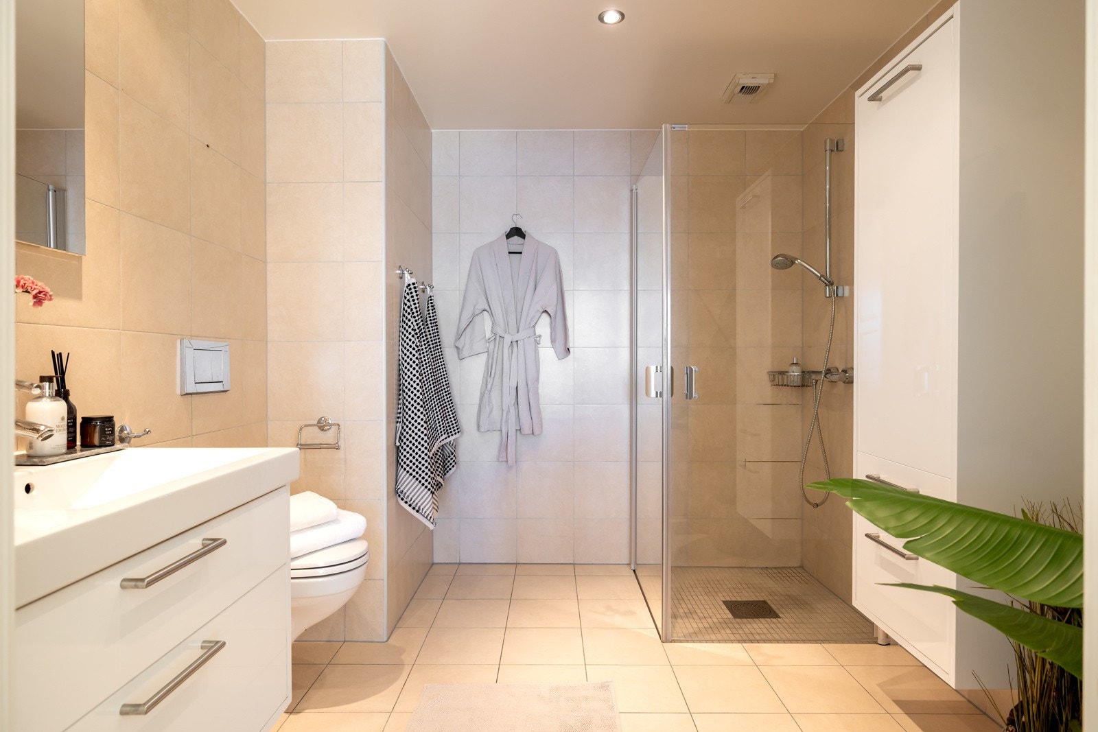 Her er det downlights i himlingen, lysbeige fliser på veggene og matchende fliser på gulv med elektrisk gulvvarme. Videre har badet et vegghengt toalett, opplegg for vaskemaskin og et dusjhjørne med innfellbare glassdører. Galleribilde