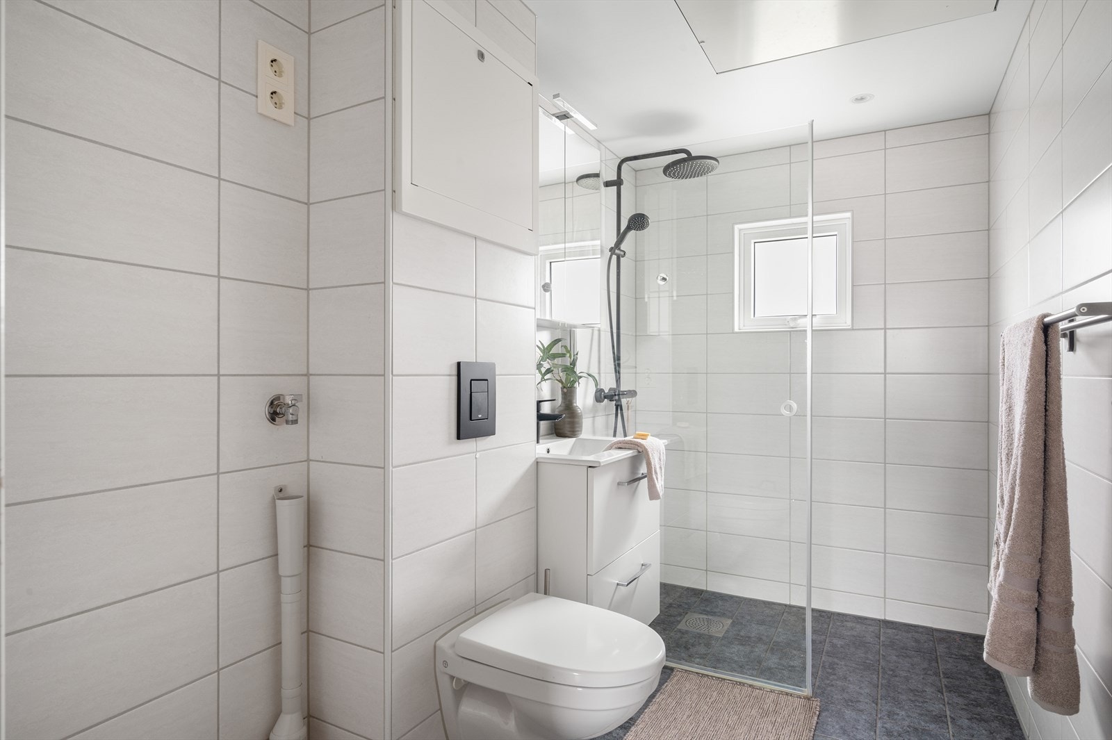 Lyst og helfliset bad utstyrt med vegghengt toalett, helstøpt servant med skuffer, dusjvegg i klart glass, opplegg for vaskemaskin og fordelerskap til rørsystemet Galleribilde