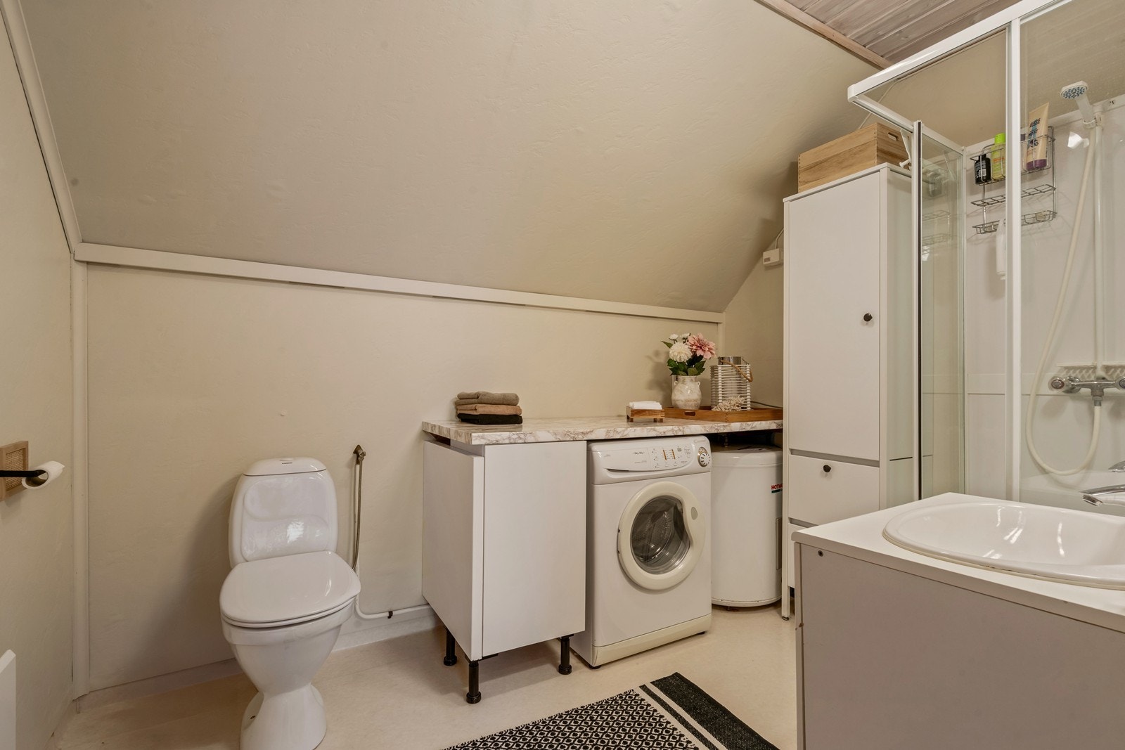 Bad/vaskerom. Veggene har våtromstapet/belegg. Taket har panel. Gulvet har vinylbelegg. Badet har dusjkabinett, seksjon med vask, toalett,
varmtvannsbereder og opplegg for vaskemaskin Galleribilde