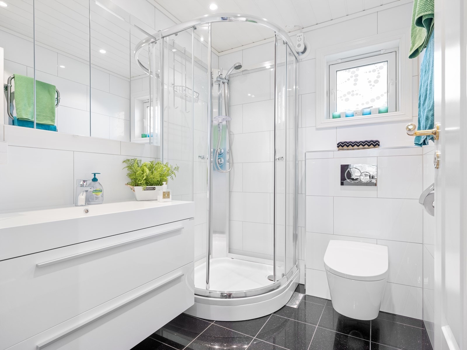 Moderne bad med fliser på gulv, vask i skuffeseksjon, dusjkabinett og vegghengt wc. Speil med belysning og spotter i tak. Galleribilde