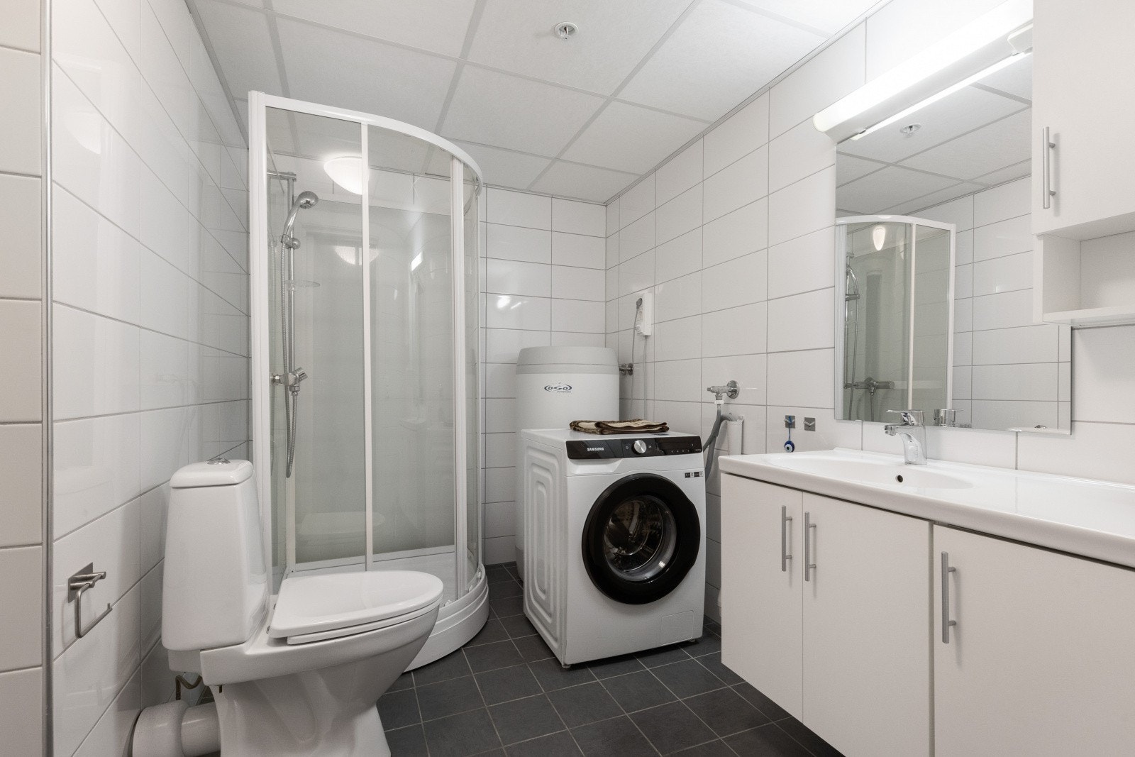 Baderom bestående av servant med underskap, gulvstående toalett, dusjkabinett, opplegg for vaskemaskin og varme i gulv Galleribilde