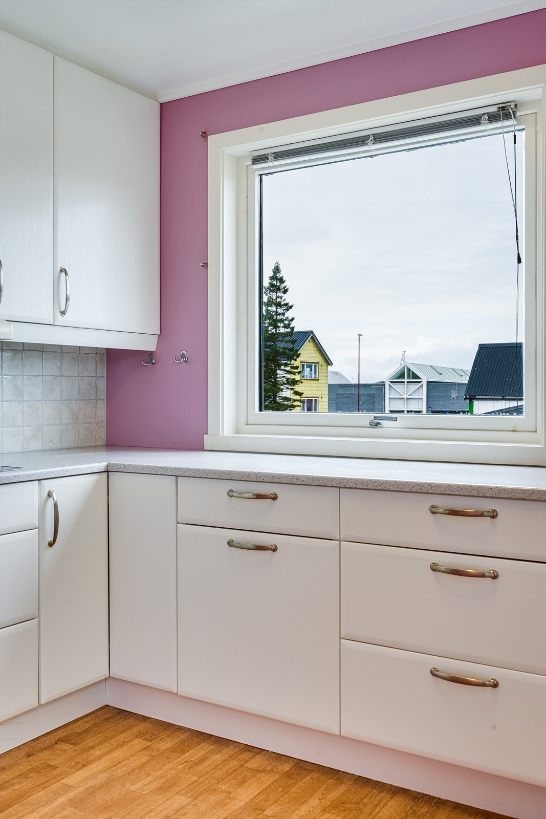 Det er fliser på vegg bak vask og platetopp, og vindu over benk med utsikt mot vest. Galleribilde