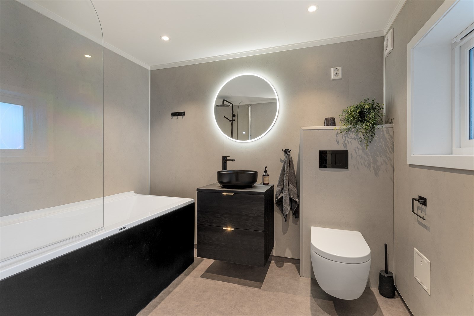 Bad frå 2021 med baderomsplater på veggar, våtromsbelegg på golv og malte plater i himling med downlights. Badet er utstyrt med vegghengt toalett, servant med innredning og badekar. Badet er også utstyrt med varmekabler i golv. Galleribilde