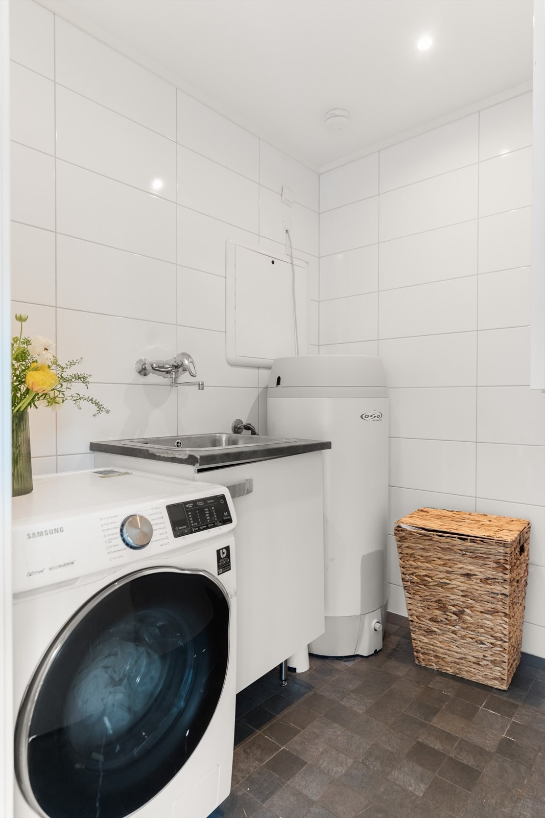 Vegg i vegg med badet er vaskerommet. Rommet har flislagte vegger og gulv. Det er videre utstyrt med stål utslagskum, opplegg for vaskemaskin, varmtvannsbereder, vannfordelerskap og stoppekran. Galleribilde