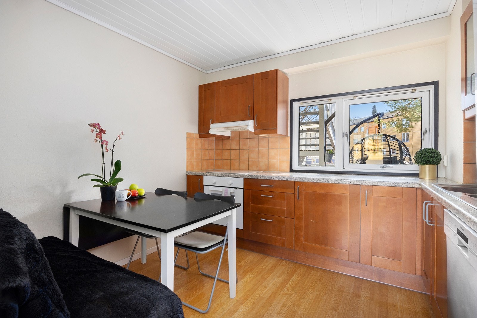 Kjøkken med lyse vegger, parkettgulv, skapfronter i tre, spiseplass og vindu som vender mot inngangspartiet. Galleribilde