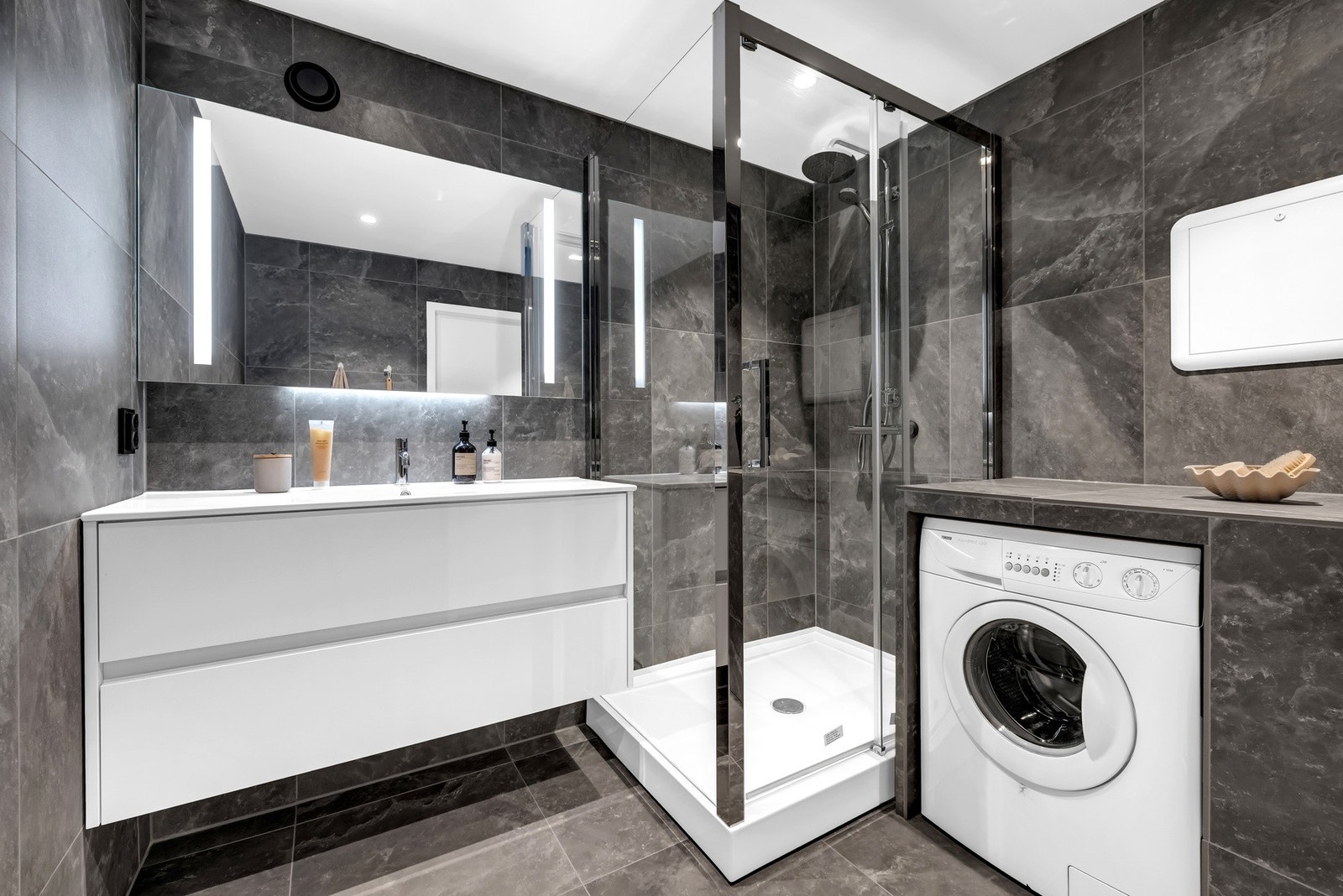 Baderommet ble totalrenovert i 2024 med store mørke fliser og flere smarte løsninger som plassbygget benk til oppvaskmaskin og dusjkabinett. Galleribilde