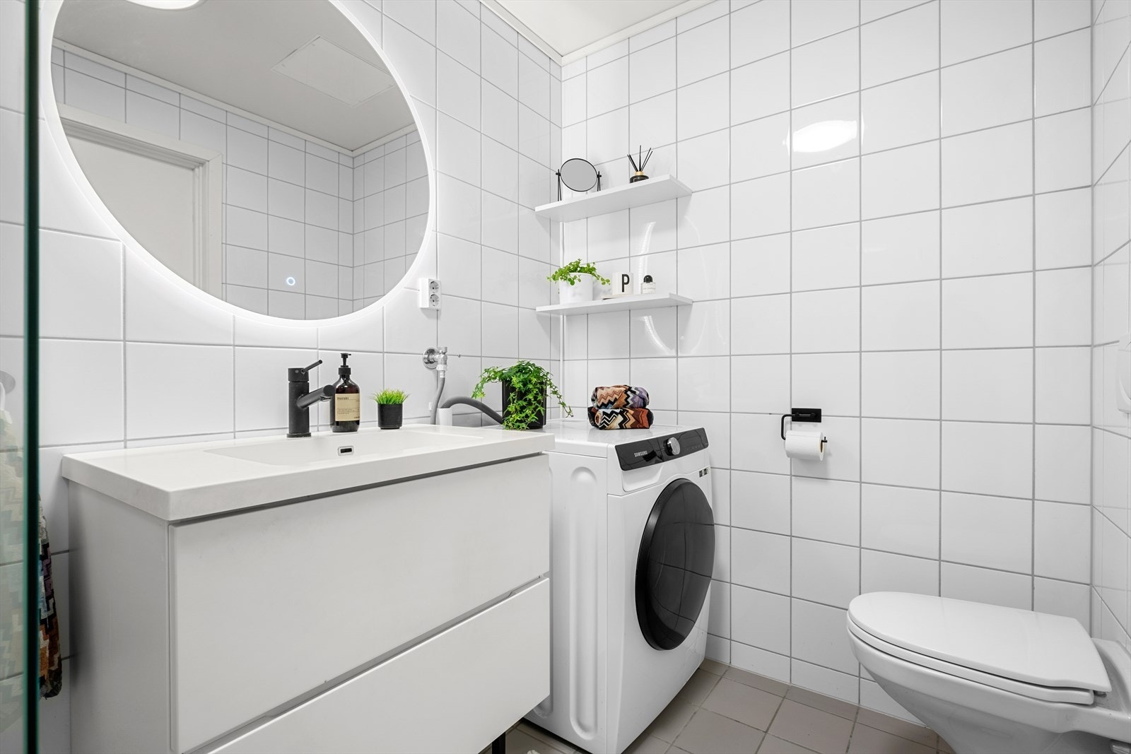 Badet er innredet med dusjvegger av glass og dusjgarnityr på vegg, veggmontert toalett, innredning med servant, opplegg for vaskemaskin (Kran og avløp) som medfølger leiligheten. Galleribilde