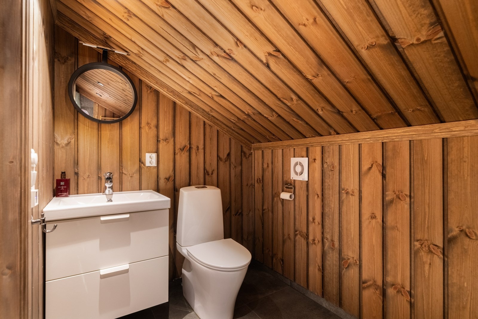 Badet er innredet med servantmøbel, toalett, dusjvegger/hjørne og opplegg for vaskemaskin. Det er elektrisk vifte på vegg. Galleribilde