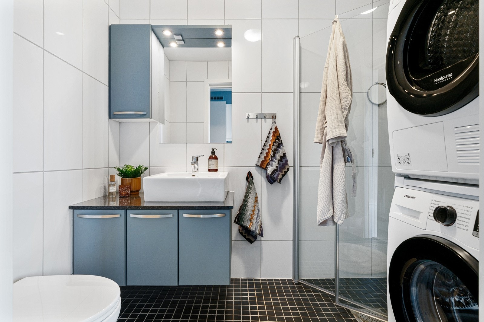 Badet er flislagt med behagelig gulvvarme. Innredningen består av overliggende servantskål, skap til oppbevaring, vegghengt toalett, dusj med innfellbare dører og opplegg for vaskemaskin. Galleribilde