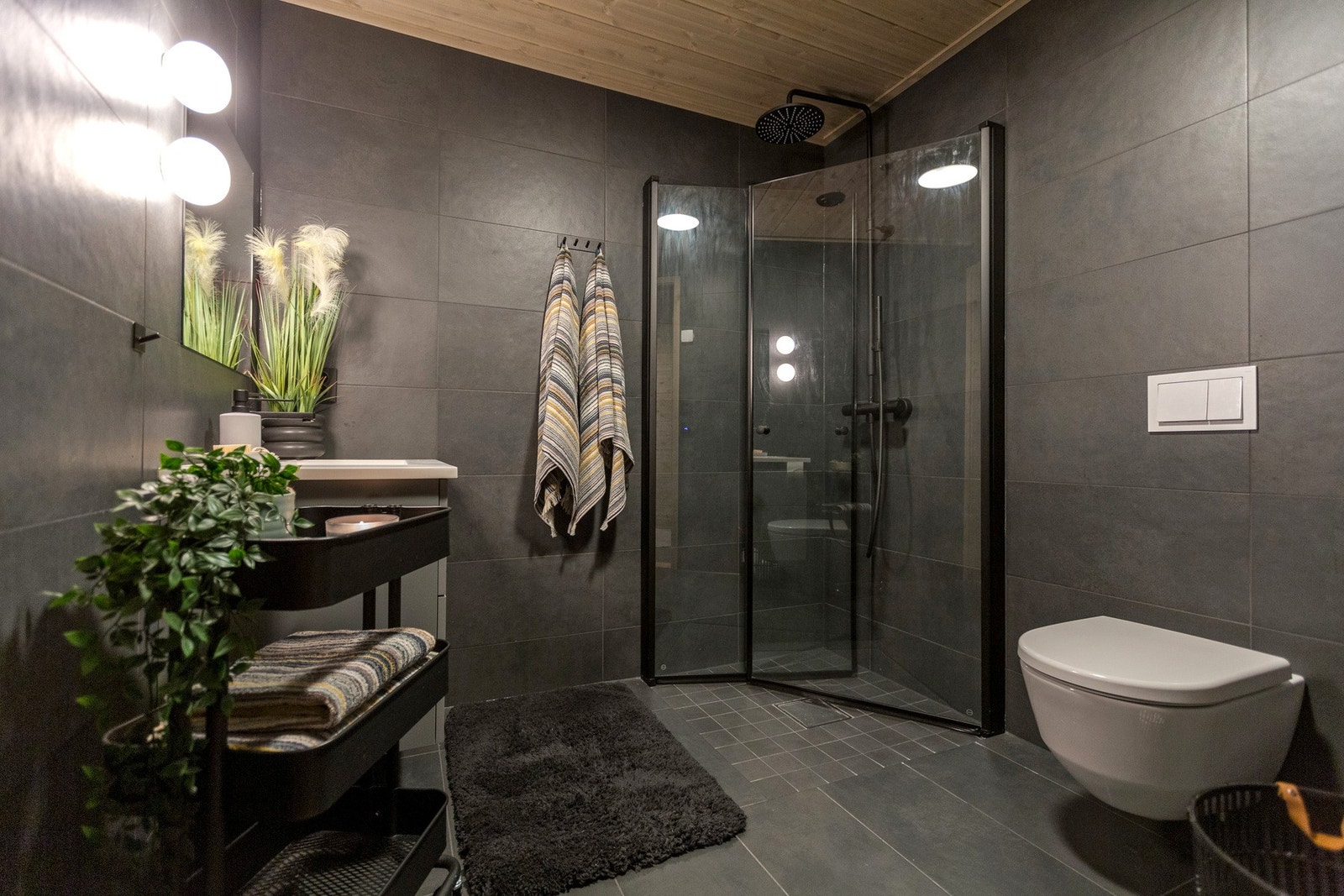 Moderne bad med mørke fliser på vegger og gulv. Romslig dusjhjørne med glassvegger og regndusj. Vegghengt toalett og servant med speil over. Tretak gir en varm kontrast til de mørke flisene. Galleribilde