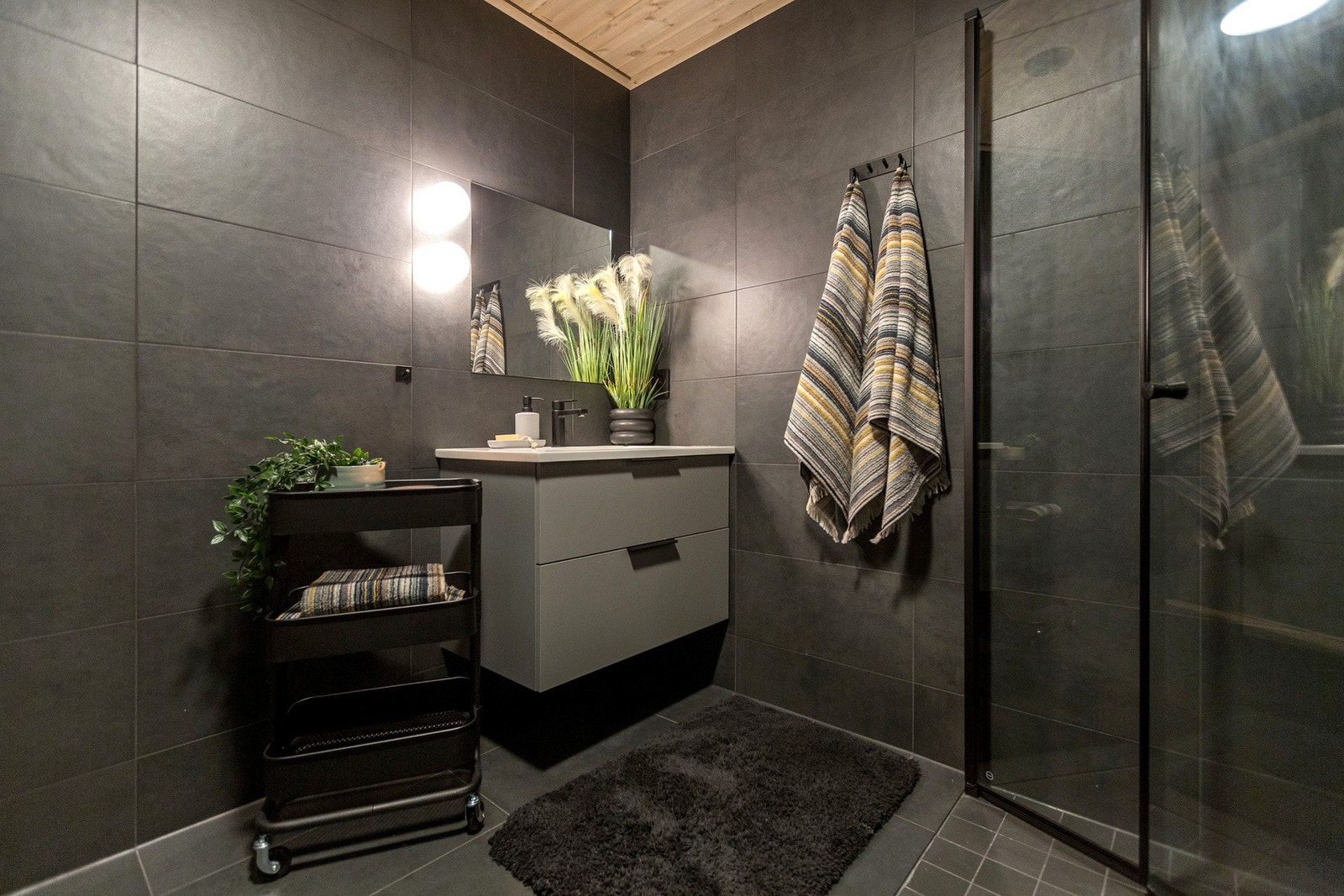 Moderne baderom med mørke fliser og dusjkabinett. Vegghengt servantskap med speil og lys. Galleribilde