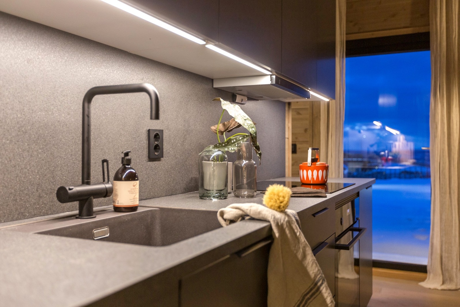 Kjøkken med mørke flater, integrert belysning under skap, og moderne armatur. Store vinduer gir godt lysinnslipp. Galleribilde