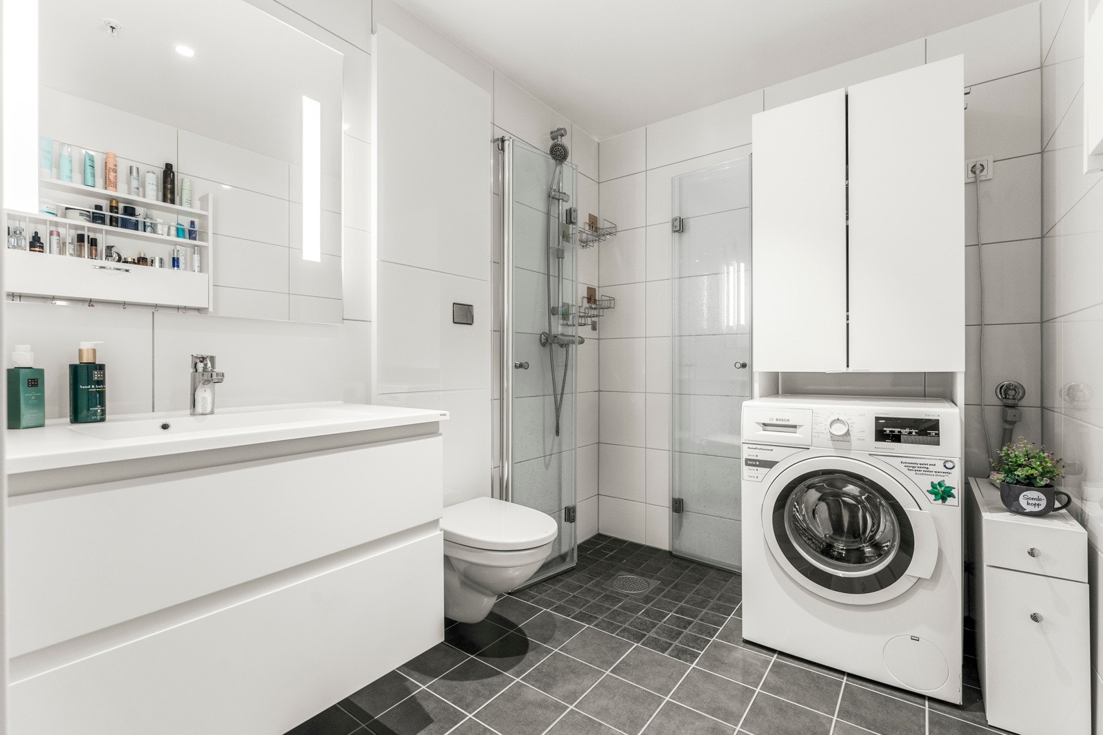 Baderommet ligger i tilknytning til boligens entré. Badet er utstyrt med dusjhjørne med innfellbare glassvegger, ett-grepsarmatur, godt med oppbevaringsplass i servantskap med heldekkende servant og ekstra belysning i speil over servanten Galleribilde