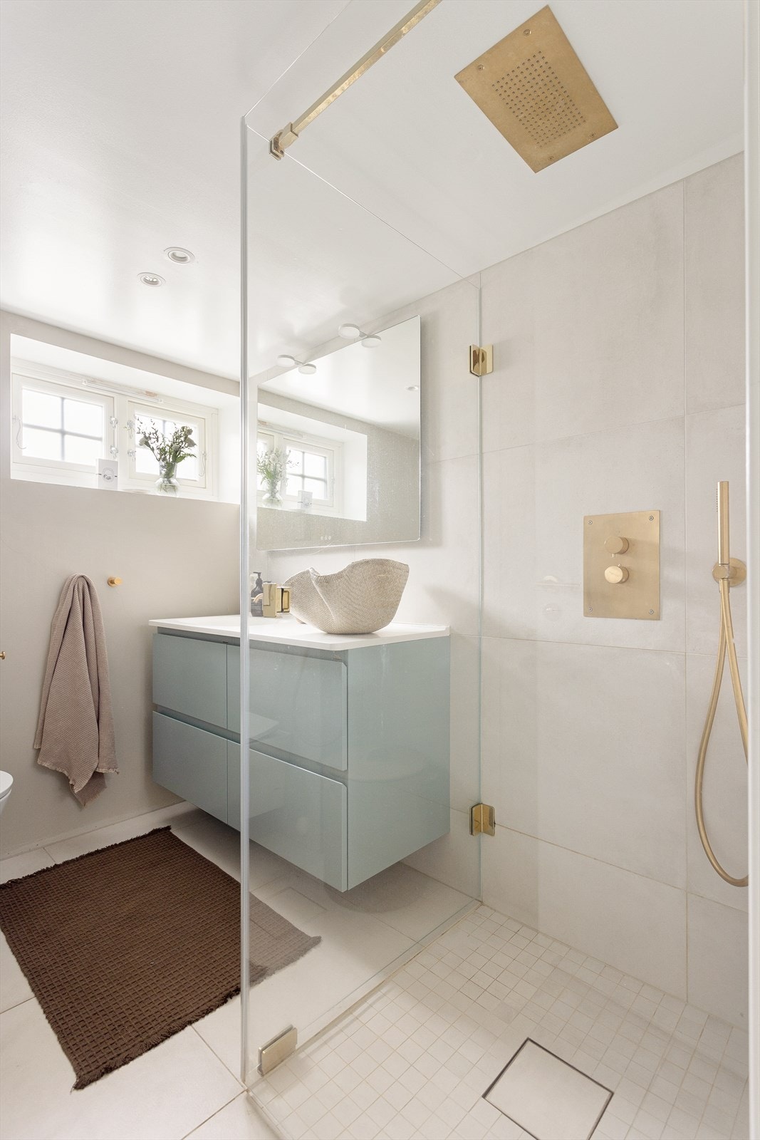 Badet er utstyrt med vegghengt wc, servant med heldekkende platetopp og skuffer under, samt dusj i hjørne med glass vegg/dør. Det er integrert dusjarmatur i børstet gull. Galleribilde