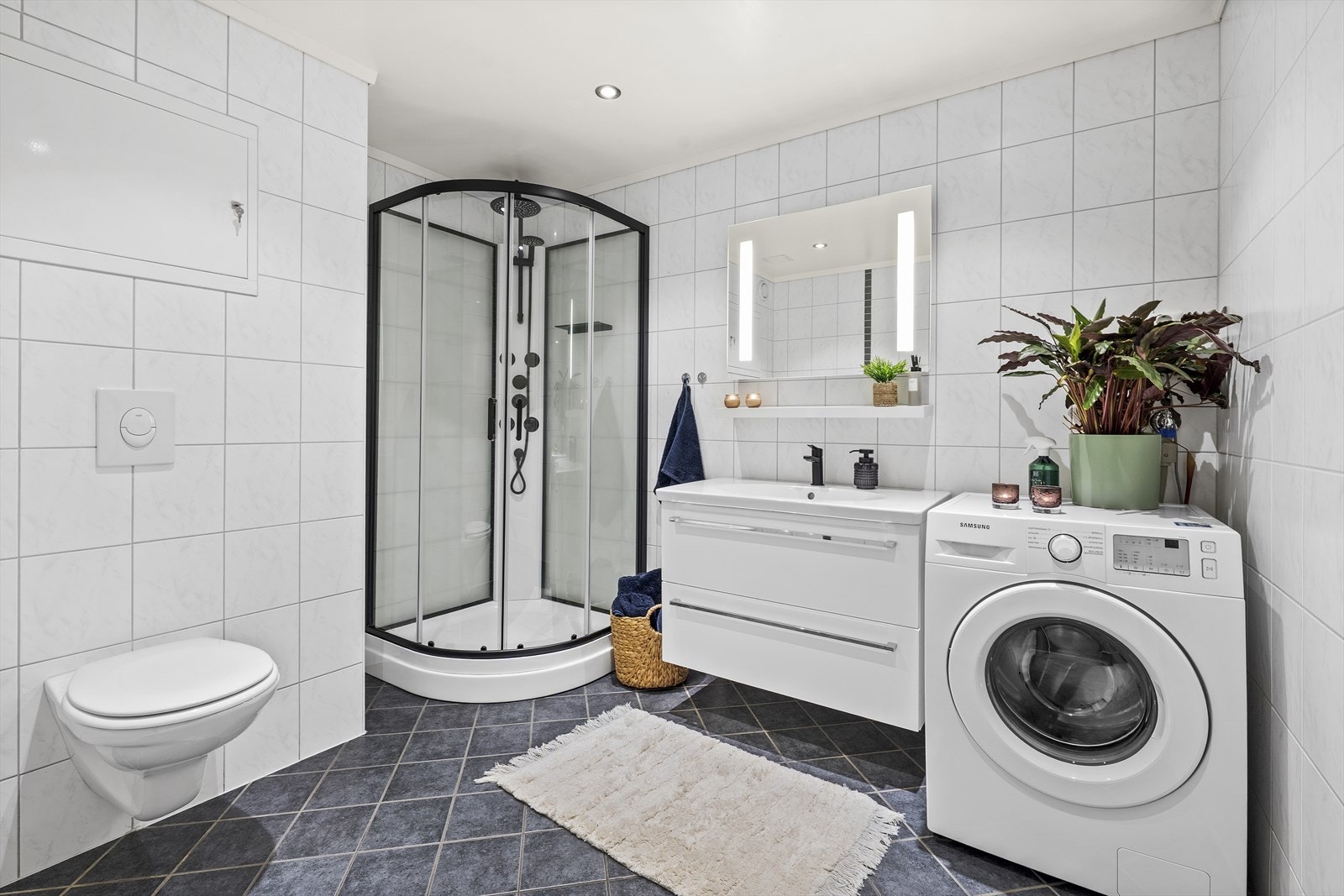 Romslig, helfliset baderom med lyse fliser på vegg og mørke fliser på gulv. Badet er utstyrt med varmekabler i gulv, heldekkende servant over skuffeinnredning, speil med belysning, dusjkabinett (2023), vegghengt toalett og opplegg til vaskemaskin Galleribilde