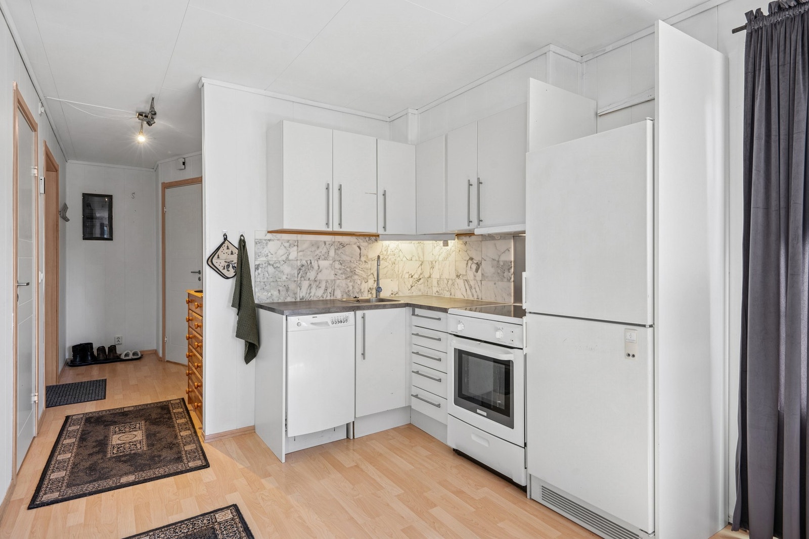 Kjøkken med åpen løsning mot stue. Innredningen består av hvite fronter, benkeplate med fliser over, og plass til oppvaskmaskin, komfyr og kjøleskap. Galleribilde