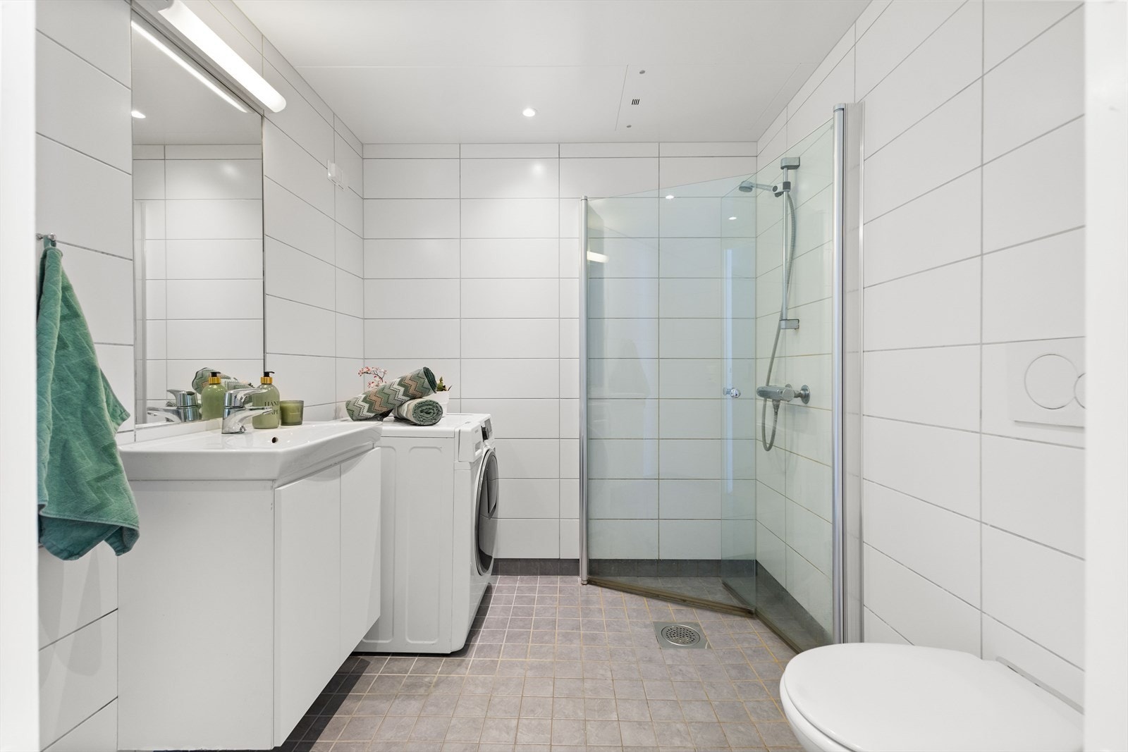 Pent flislagt baderom med varmekabler i gulv, opplegg for vaskemaskin, dusjhjørne med innfellbare dusjdører, toalett og servant med innredning Galleribilde