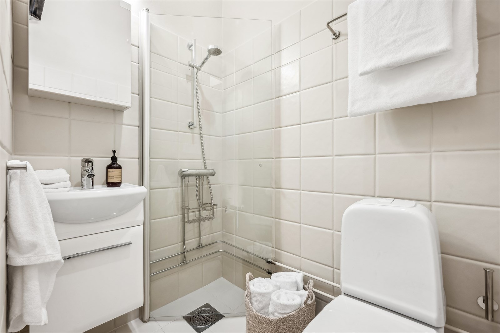 Pent baderom med lysmalte fliser. Innredning består av dusj med glassvegg og innredning med heldekkende servant og toalett. Galleribilde