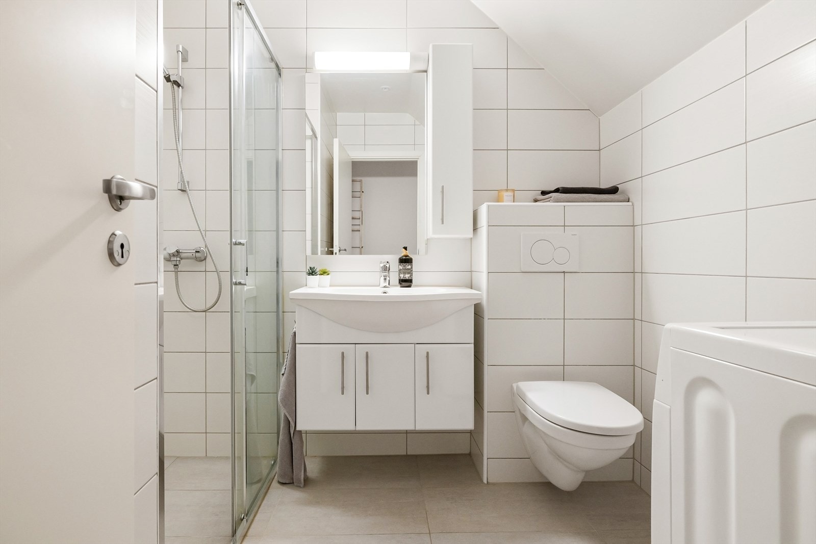 Badet er helfliset og inneholder dusj, servant med over- og underskap, vegghengt toalett og opplegg for vaskemaskin. Galleribilde