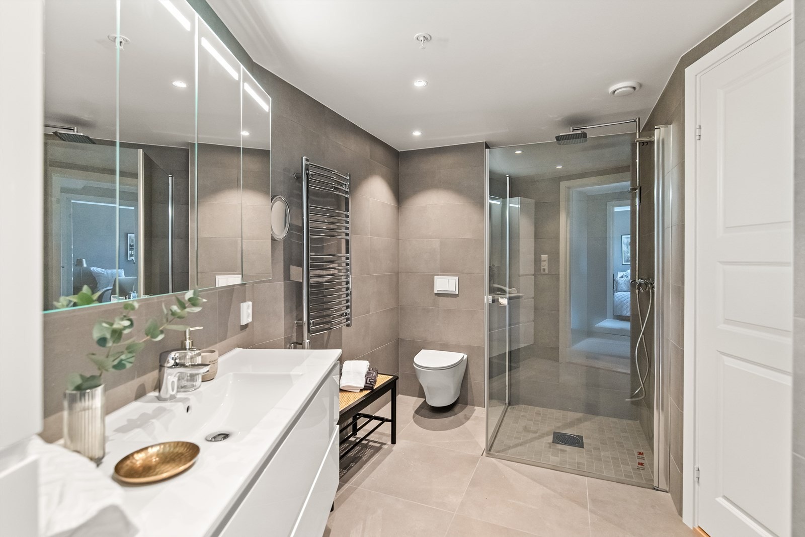 Planløsningen byr på et moderne bad med tilliggende vaskerom. Alle gulv og vegger er kledd med store, grå fliser for et stilrent uttrykk. Downlights og gulvvarme er installert, og badet har elektrisk håndklevarmer og et sminkespeil med tilhørende lys. Galleribilde