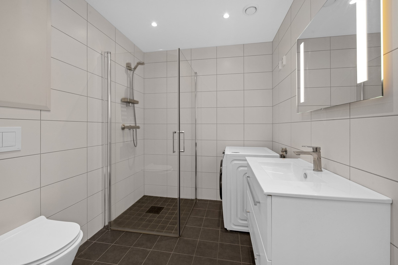 Delikat bad med dusj, vegghengt toalett, vask i pen innredning og speil med lys innfelt. Det er også plass/opplegg for vaskemaskin på badet Galleribilde