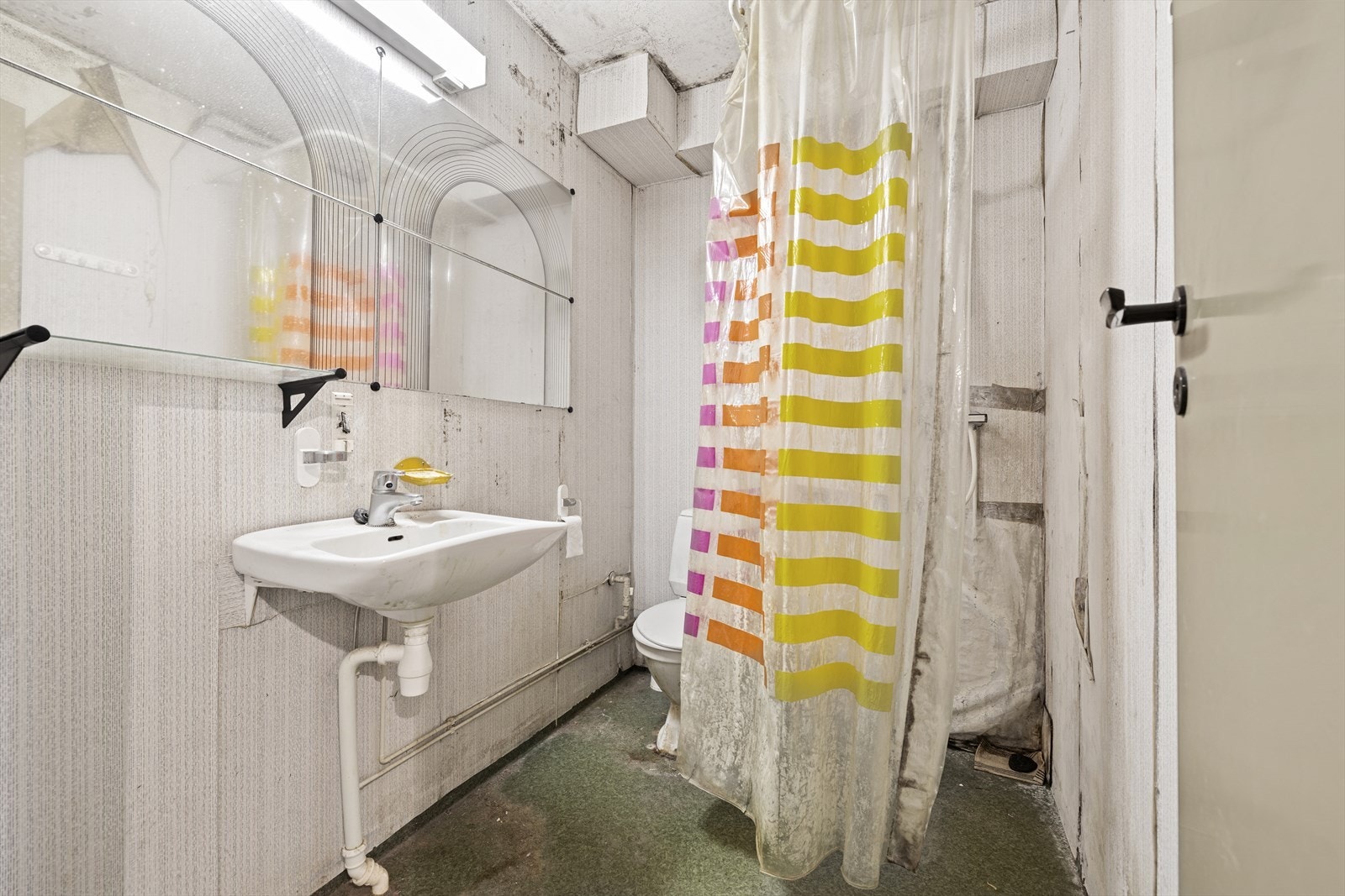 Badet er enkelt innredet med dusjhjørne, gulvstående toalett og en enkel servant. Rommet fremstår med betydelig slitasje og har behov for totalrenovering. Galleribilde