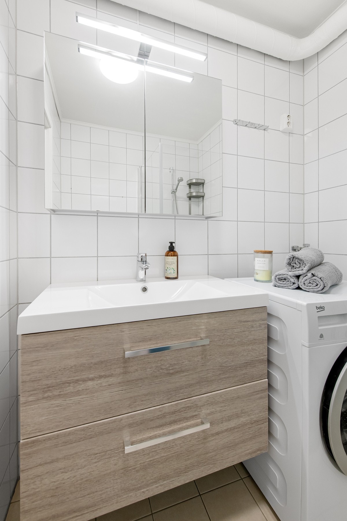 Flislagte overflater og gulv med gulvvarme gir et helhetlig og ryddig uttrykk. Rommet har vegghengt servantskap med skuffer, ovenpåliggende servant med ett-greps armatur og speilskap med belysning. Galleribilde